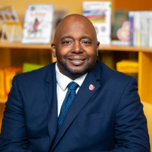 Professional headshot of Dr. Ronald White wearing a navy suit and blue tie, seated in front of bookshelves and smiling at the camera.